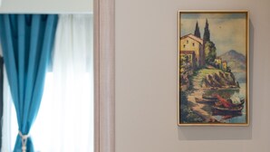 A room with a painting of a coastal scene, a white chair, a table, and a curtain. - MYKONIATIKA VILLAS (Nea Propontida)