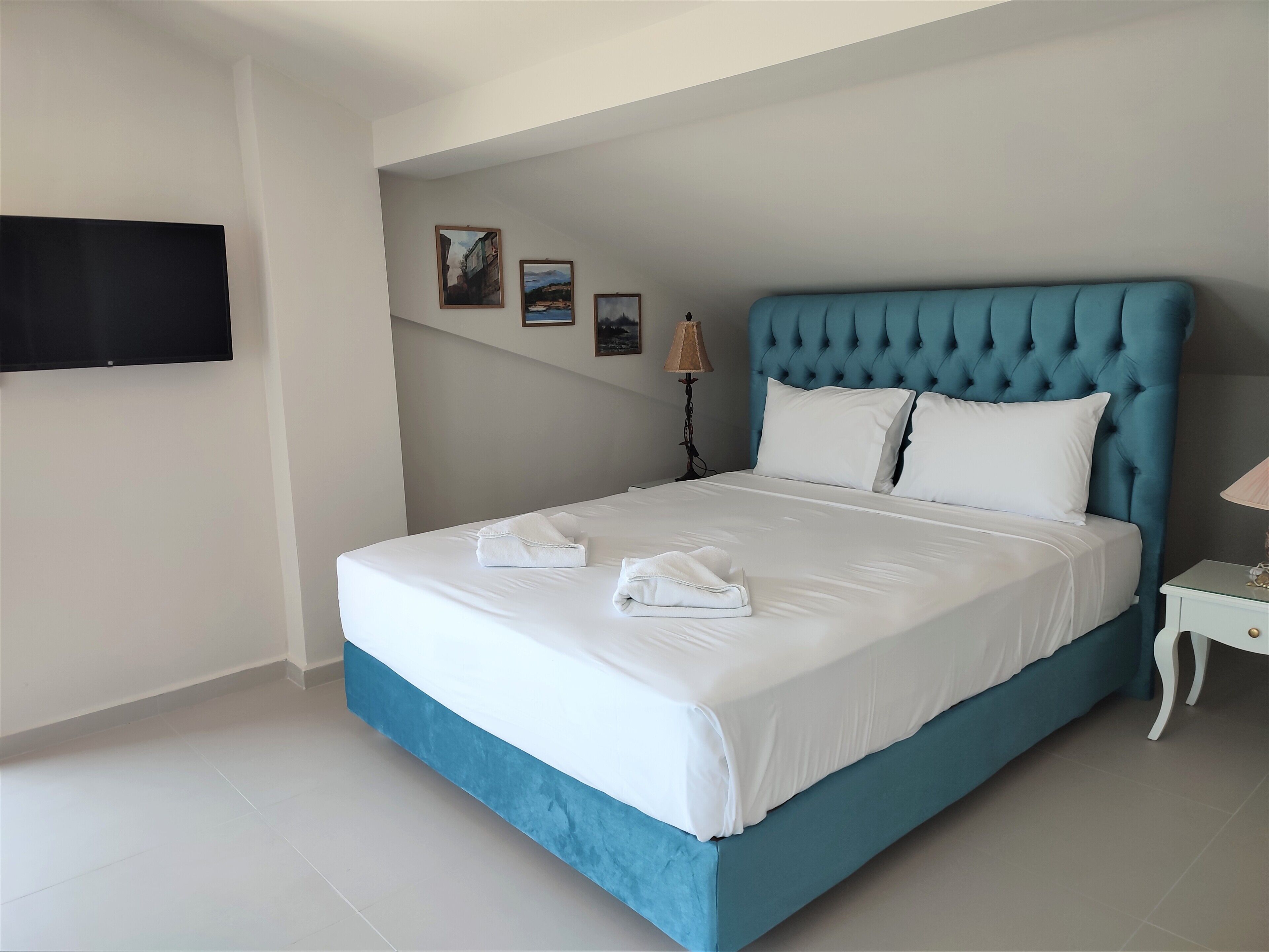 Un lit bien fait, avec du linge de lit blanc et une tête de lit bleue, un téléviseur à écran plat fixé au mur et une petite table de chevet avec une lampe.