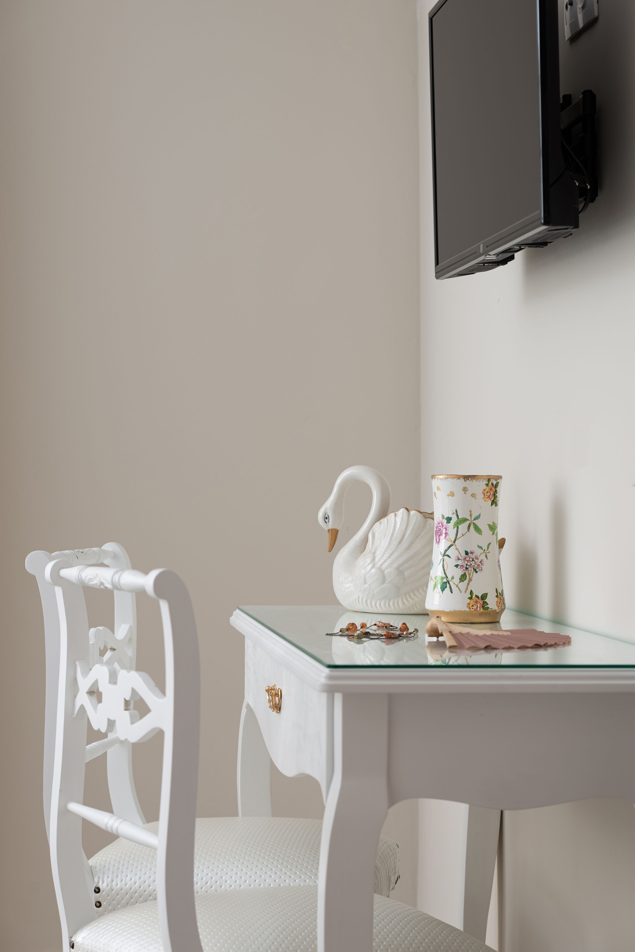 Ein weißer Stuhl mit einer verzierten Rückenlehne, ein Glastisch mit einer schwanförmigen Vase und einer tasse mit Blumenmuster, sowie ein Flachbildfernseher an der Wand.