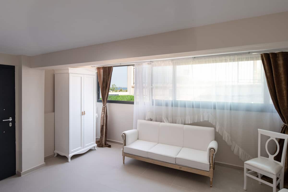 A room with a white sofa, a white chair, a wardrobe, and a window with sheer curtains.