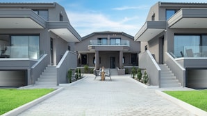 A modern residential building with a central entrance, two staircases on either side, and a paved driveway leading to a seating area. - MYKONIATIKA VILLAS (Nea Propontida)
