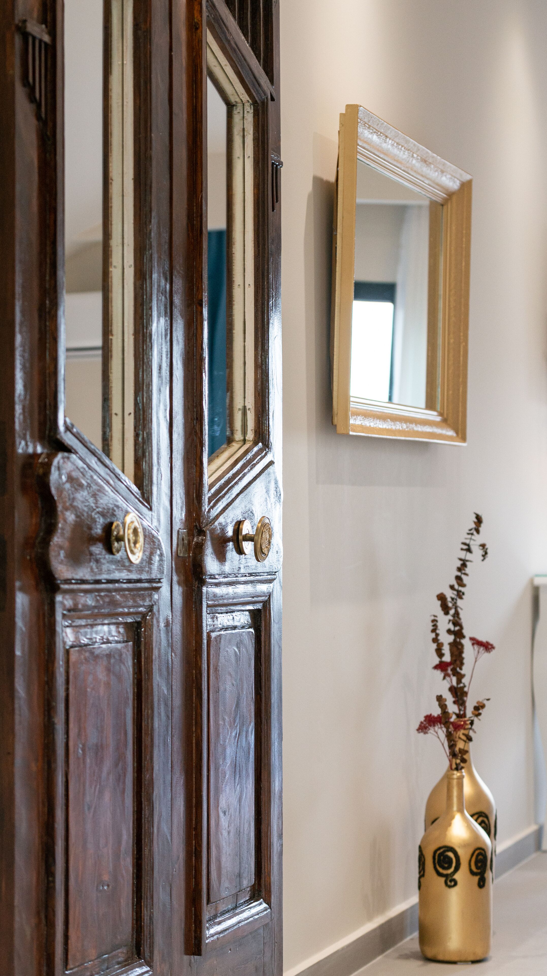 A wooden door with brass handles, a mirror on the wall, and a vase with dried flowers.