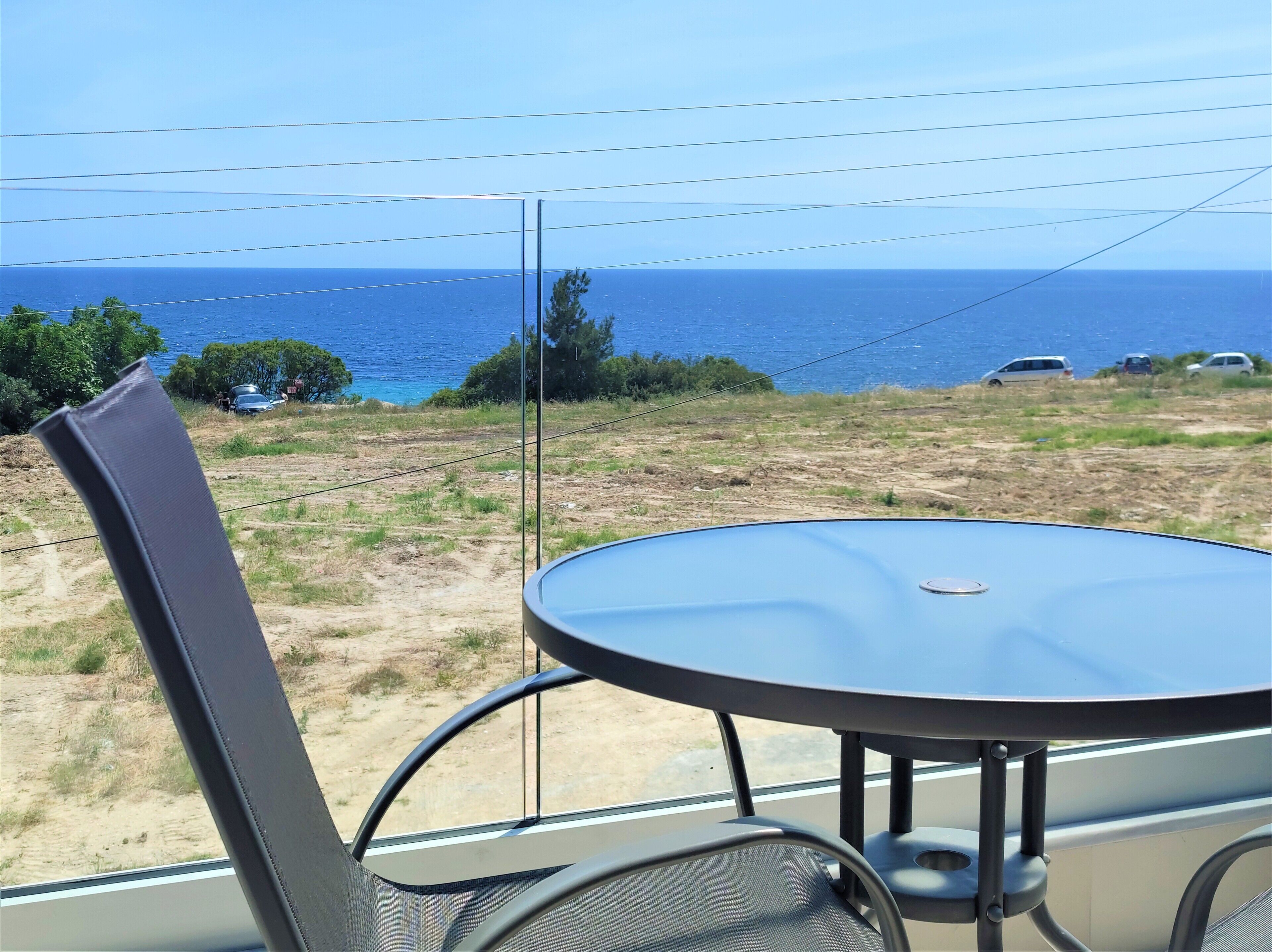 Un balcón con una mesa redonda y sillas, con vistas a un paisaje costero de vegetación escasa y coches estacionados.