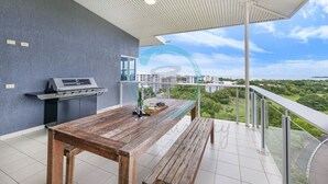 Un balcón con mesa y parrilla de madera, vista a un paisaje urbano y cielo despejado.