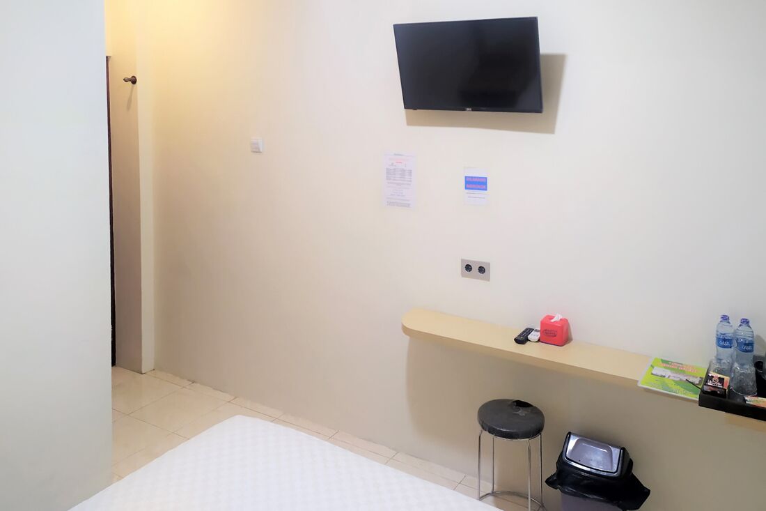 A hotel room with a flat-screen TV mounted on the wall, a small desk with a chair, a trash bin, and a shelf with bottled water and a remote control.