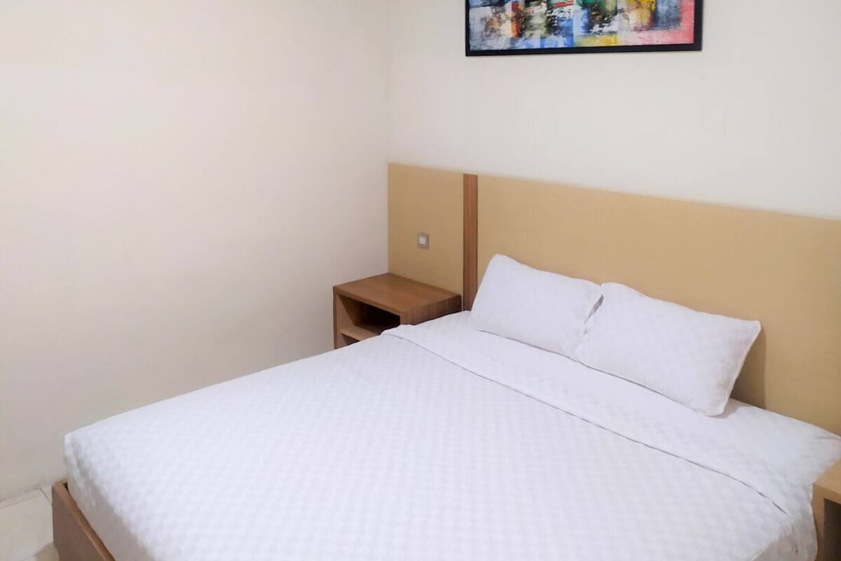 Ein ordentlich bezogenes Bett mit weißer Bettwäsche, einem Holz-Kopfteil und einem kleinen Nachttisch daneben.