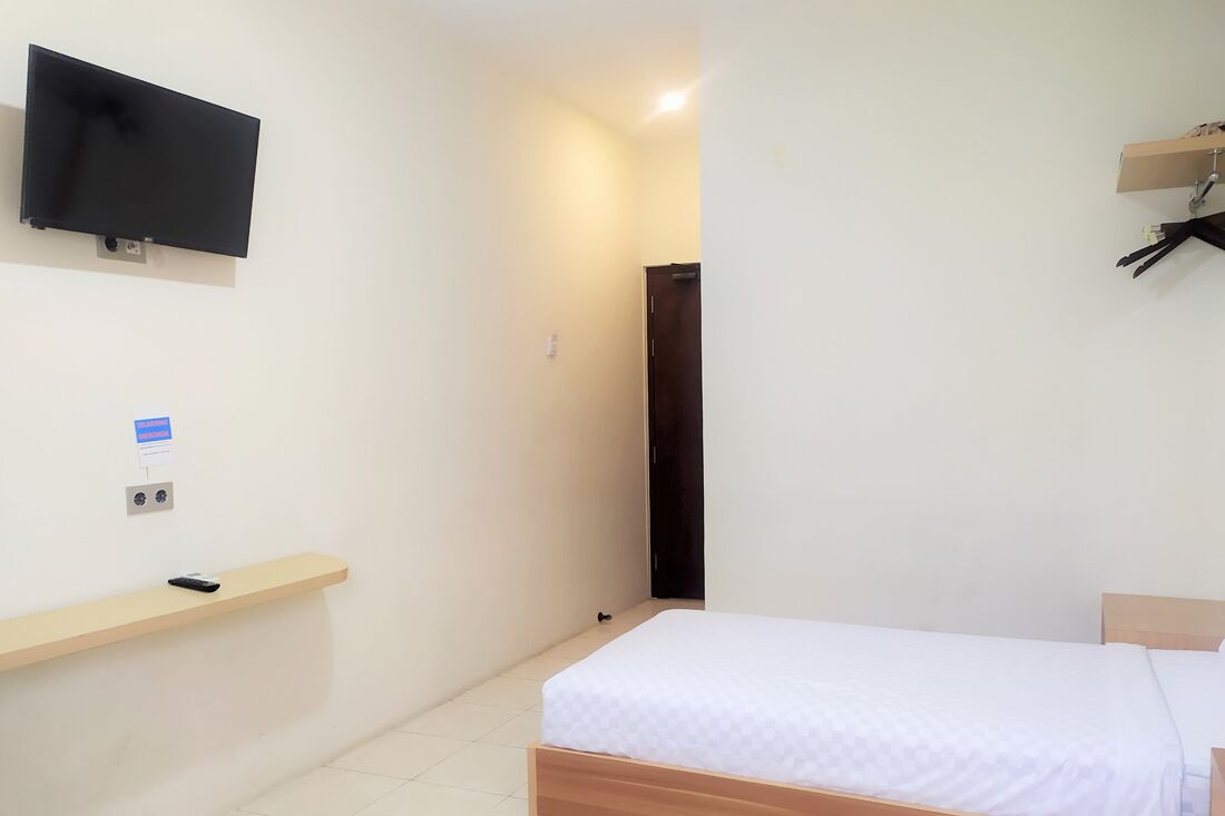 Una camera piccola e pulita con un letto, una TV a muro, uno scaffale con un telecomando e una porta che dà su un'altra stanza.