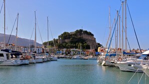 Un port de plaisance où sont amarrés de nombreux bateaux, une colline imposante sur laquelle se dresse une structure ressemblant à un château, et des bâtiments le long de la promenade.