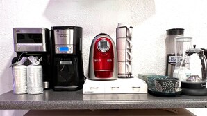 Un comptoir de cuisine avec une machine à café, un grille-pain et un mixeur. Une étagère au-dessus contient divers aliments, et un meuble en dessous, doté de portes en verre, renferme des ustensiles et des récipients de cuisine.