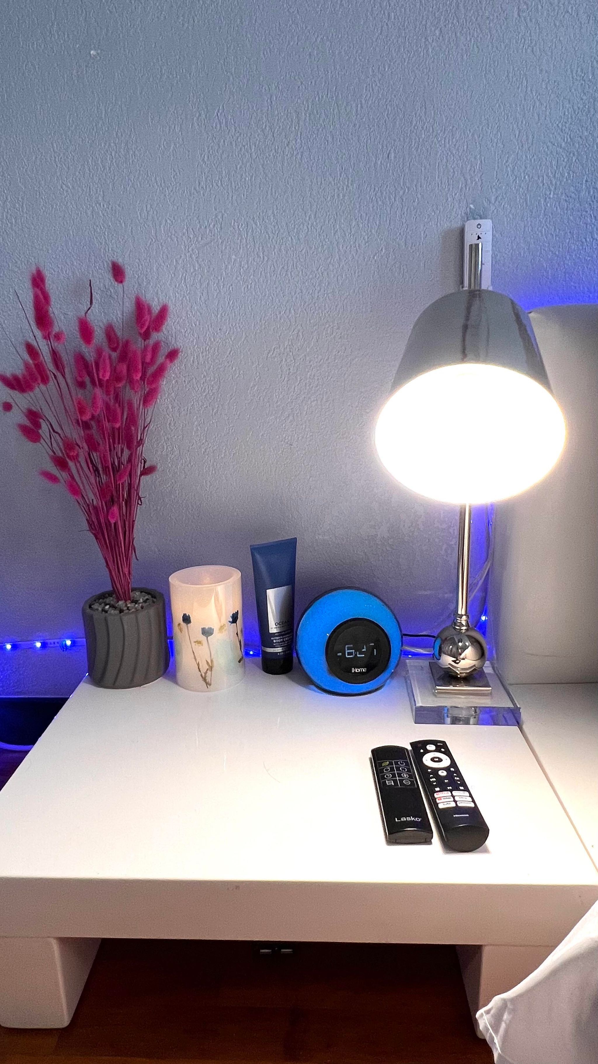 A bedside table with a lamp, a remote control, a blue clock, a small potted plant, and a candle.