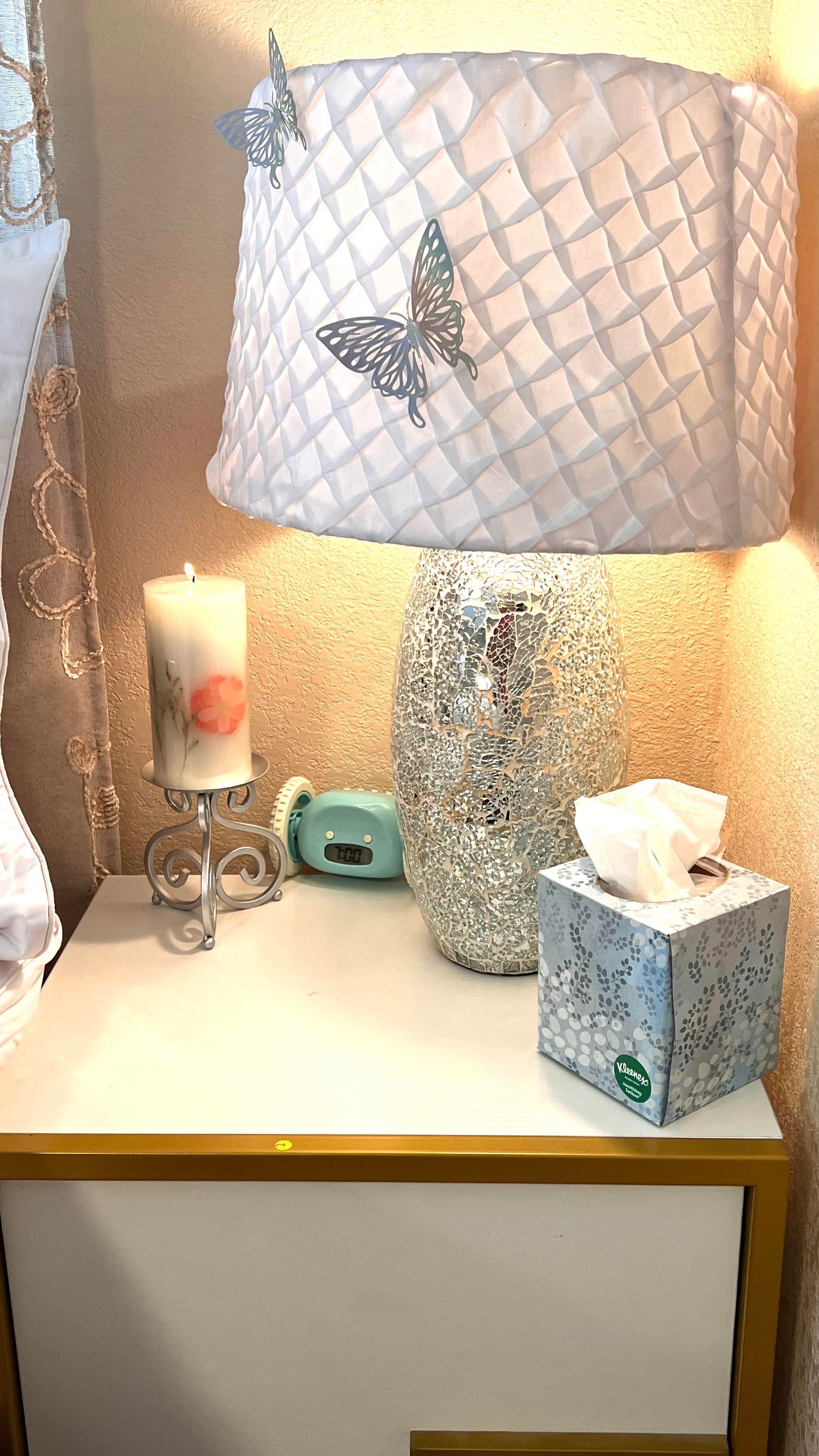 A bedside table with a white lamp featuring a butterfly design, a lit candle, a tissue box, and a clock.