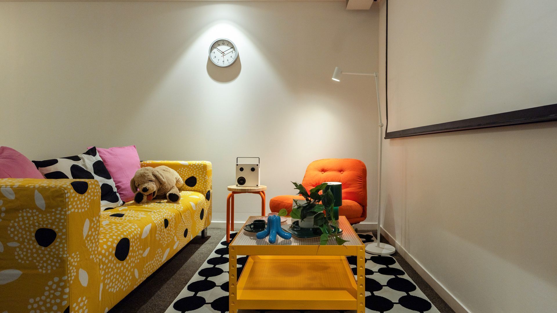 A cozy living room with a yellow sofa, a red armchair, a small table with a speaker, and a clock on the wall.