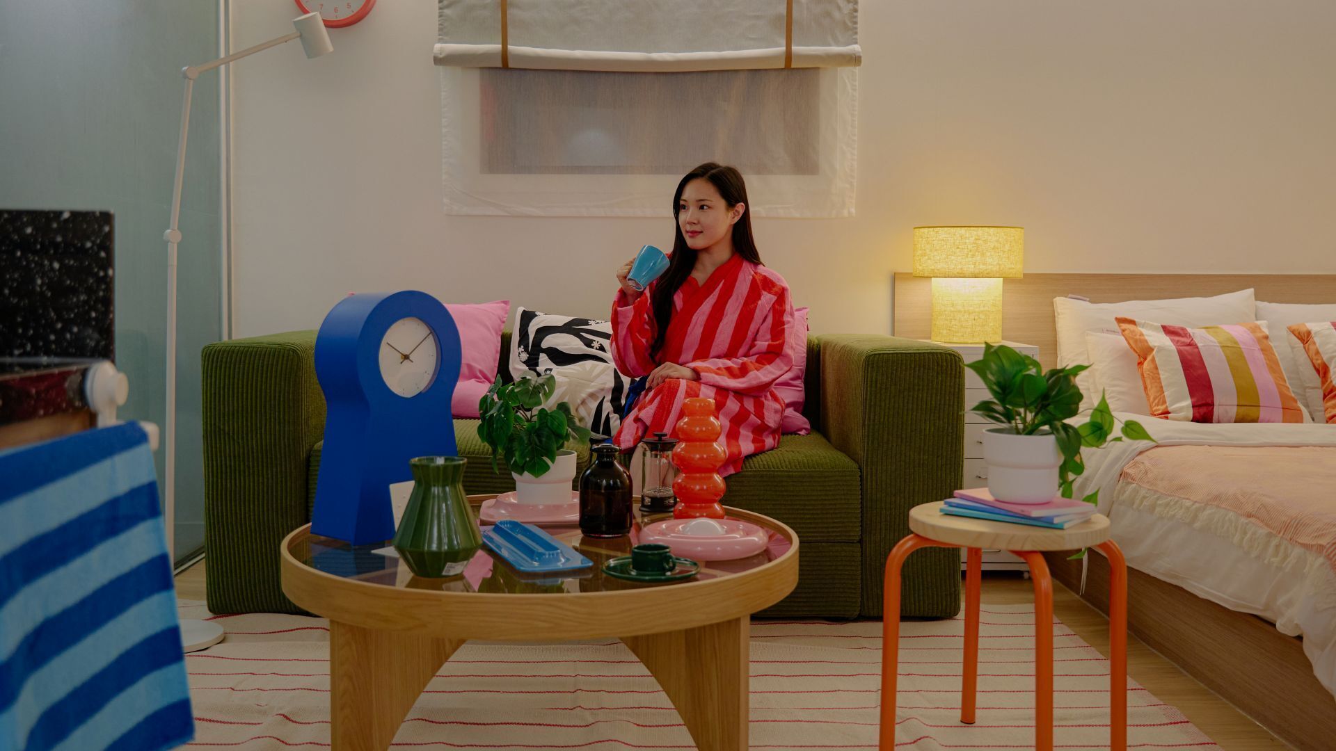 A cozy hotel room with a green sofa, a wooden coffee table with various items, a blue clock, and a bedside table with a lamp.