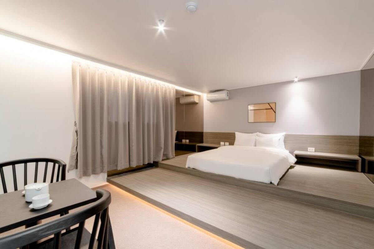 A modern hotel room with a large bed, bedside tables, a small table with chairs, and a window with curtains.