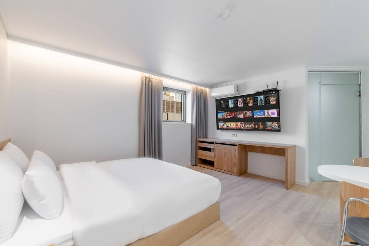 A modern hotel room with a large bed, a desk, a TV mounted on the wall displaying a movie selection, and a small seating area.