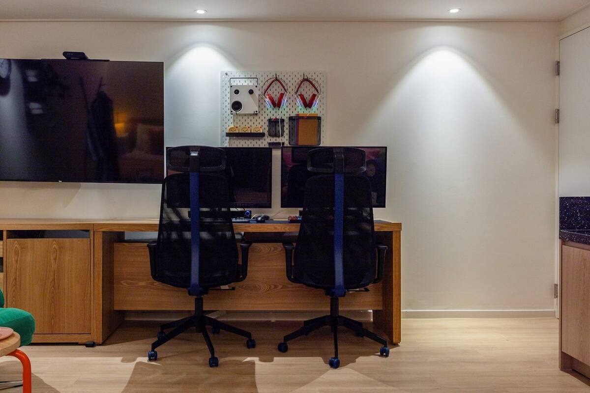 A modern office space with a large flat-screen TV mounted on the wall, two office chairs, a wooden desk, and a small table with a green chair and a pink cup.