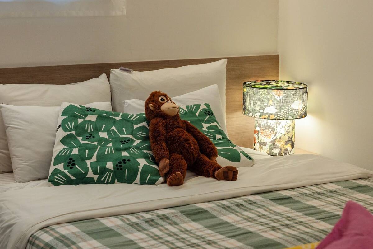 A bed with a checkered bedspread, a green leaf-patterned pillow, a brown plush toy, and a lamp with a patterned shade.
