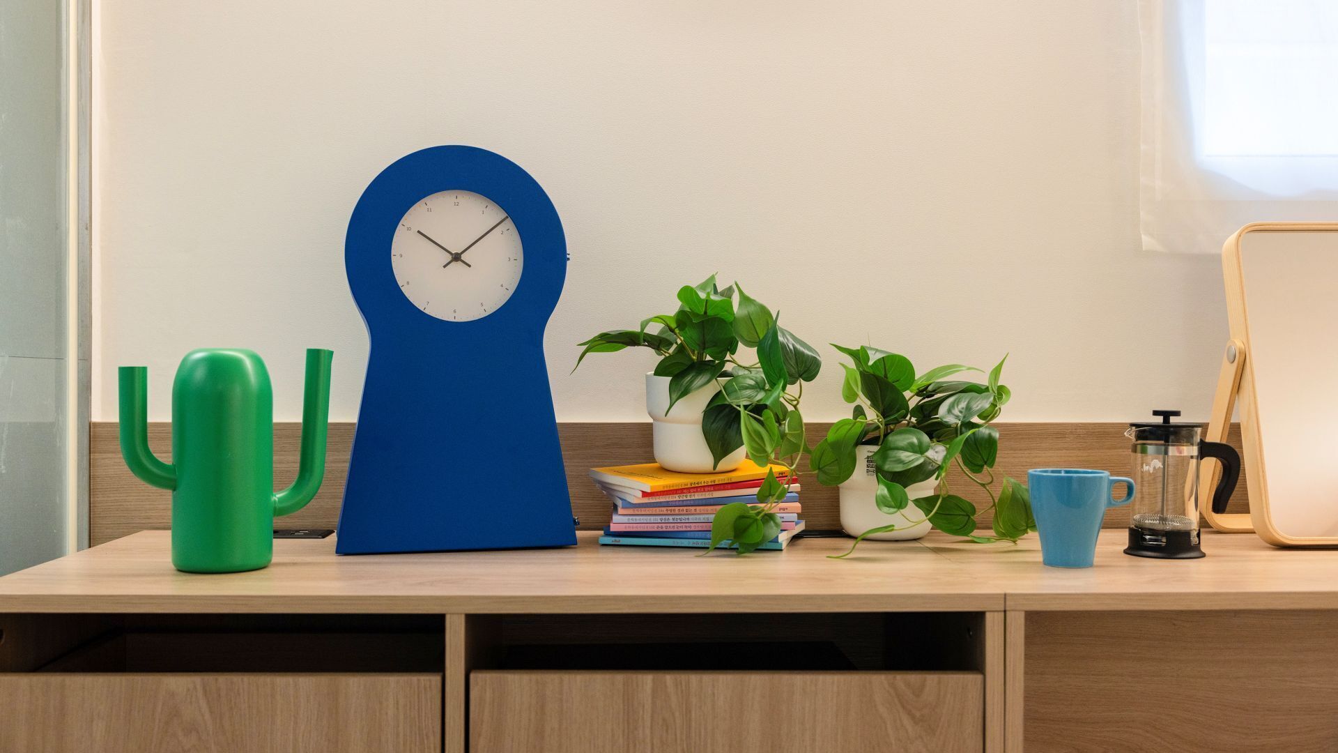A wooden desk with a blue clock, green cactus, potted plants, books, a blue mug, and a coffee maker.