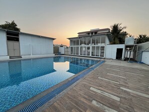 A swimming pool with a tiled deck, surrounded by buildings with large windows and a clear sky. - Hotel Jhankar Palace (Dhule)