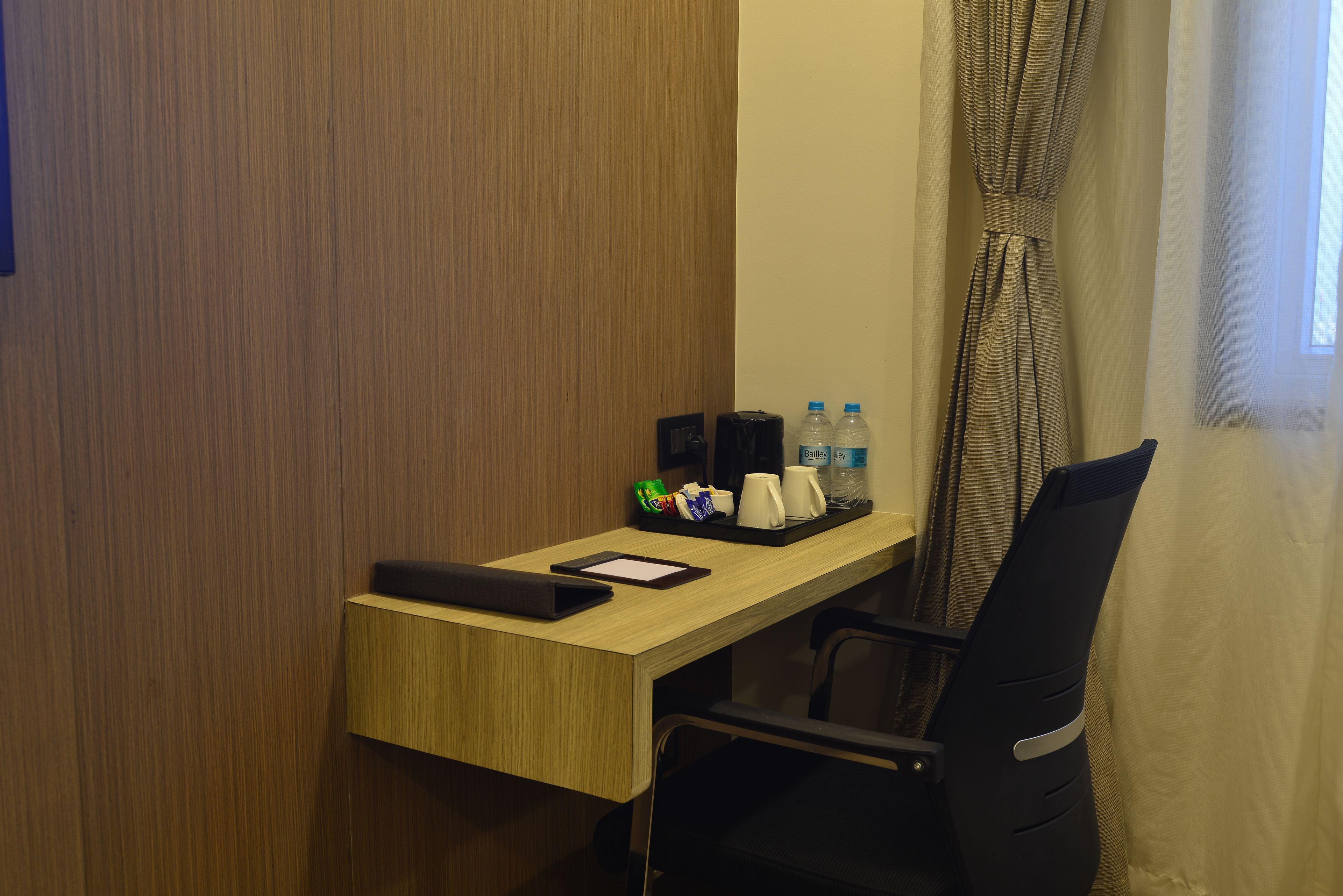 A hotel room with a wooden desk, a black chair, a black bag, a tray with a water bottle, a mug, and a small green bag.