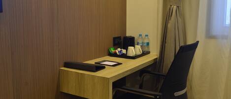 A hotel room with a wooden desk, a black chair, a black bag, a tray with a water bottle, a mug, and a small green bag.