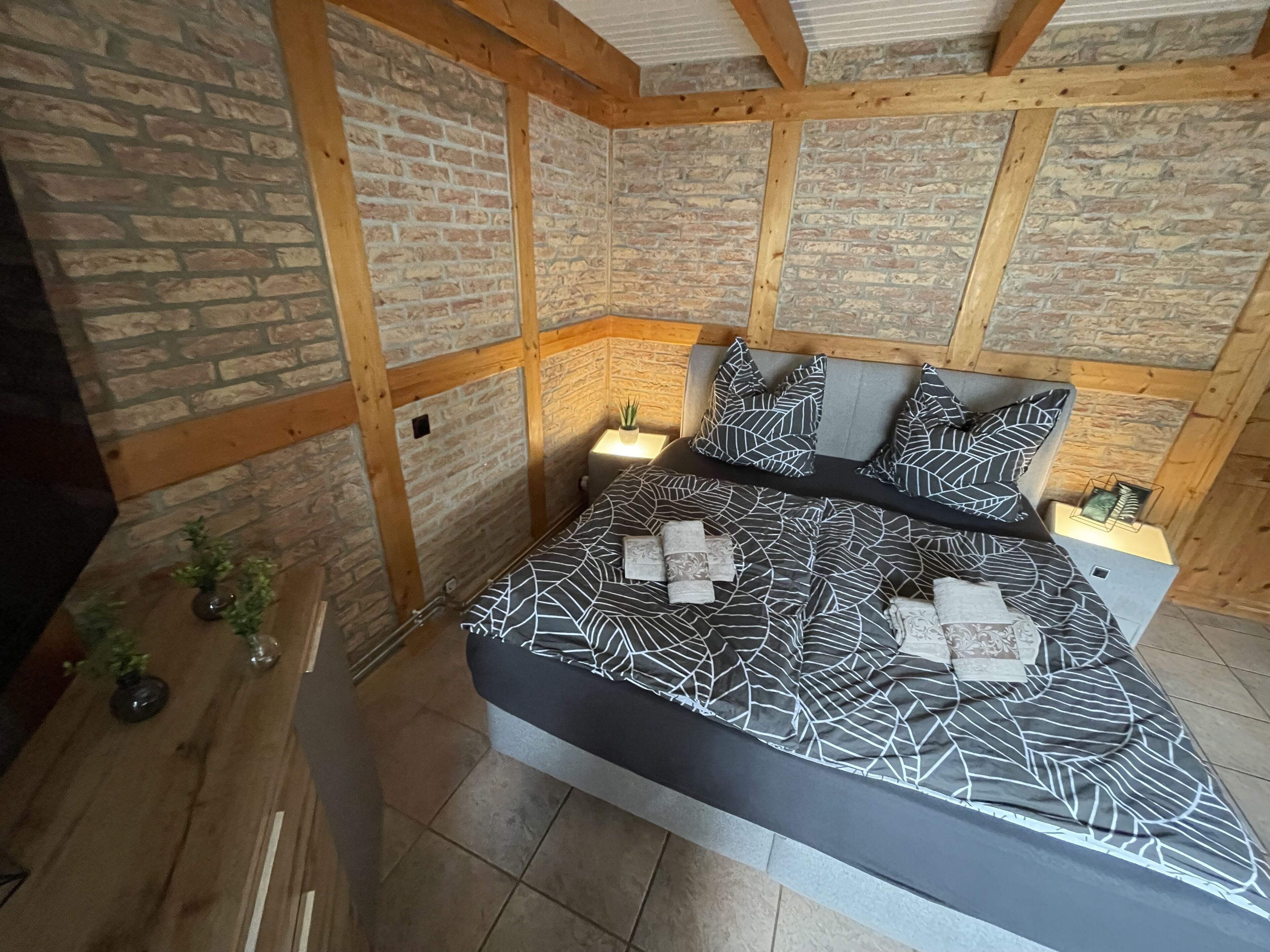 Un dormitorio con pared de piedra, vigas de madera, una cama con cubrecama estampado con motivos de hojas, dos mesitas de noche y una pequeña planta.
