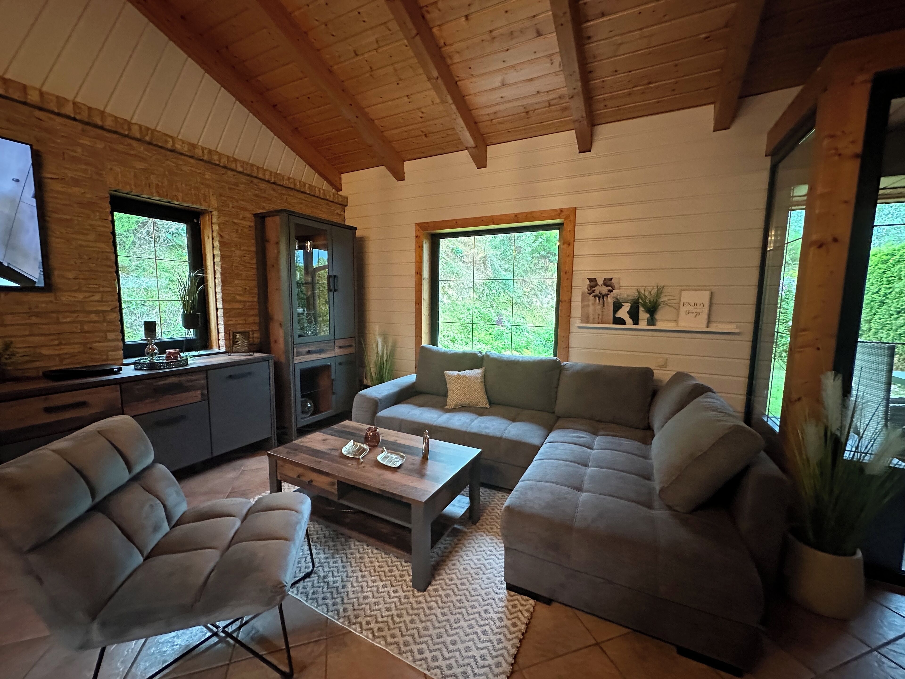 A cozy living room with a wooden ceiling, a grey sofa, a coffee table, and a TV mounted on the wall.