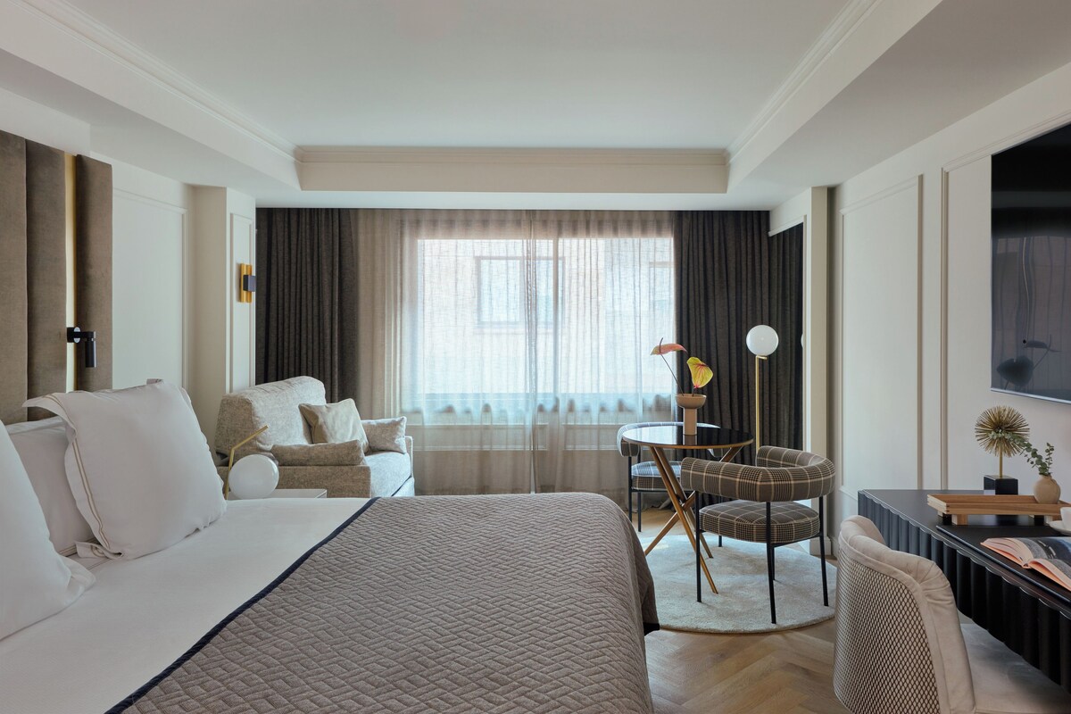 A hotel room with a large bed, a seating area with a table and chairs, a TV, and a window with curtains.