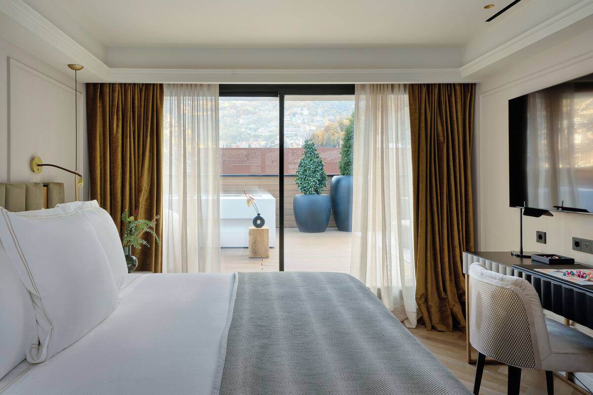 A hotel room with a large bed, a desk, a chair, a TV, and a view of the outside.