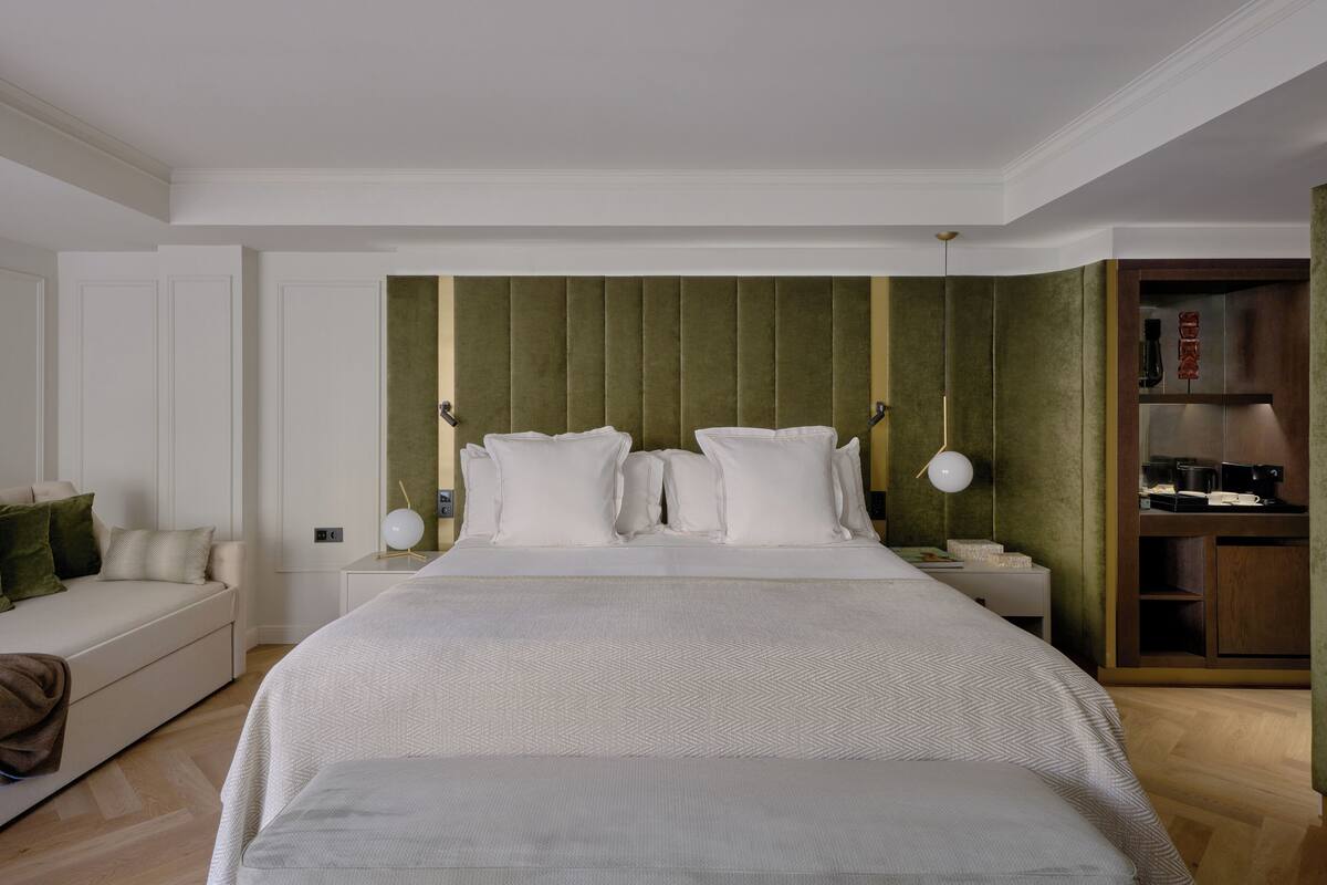 A hotel room with a large bed, a bedside table with a lamp, a sofa, and a minibar.
