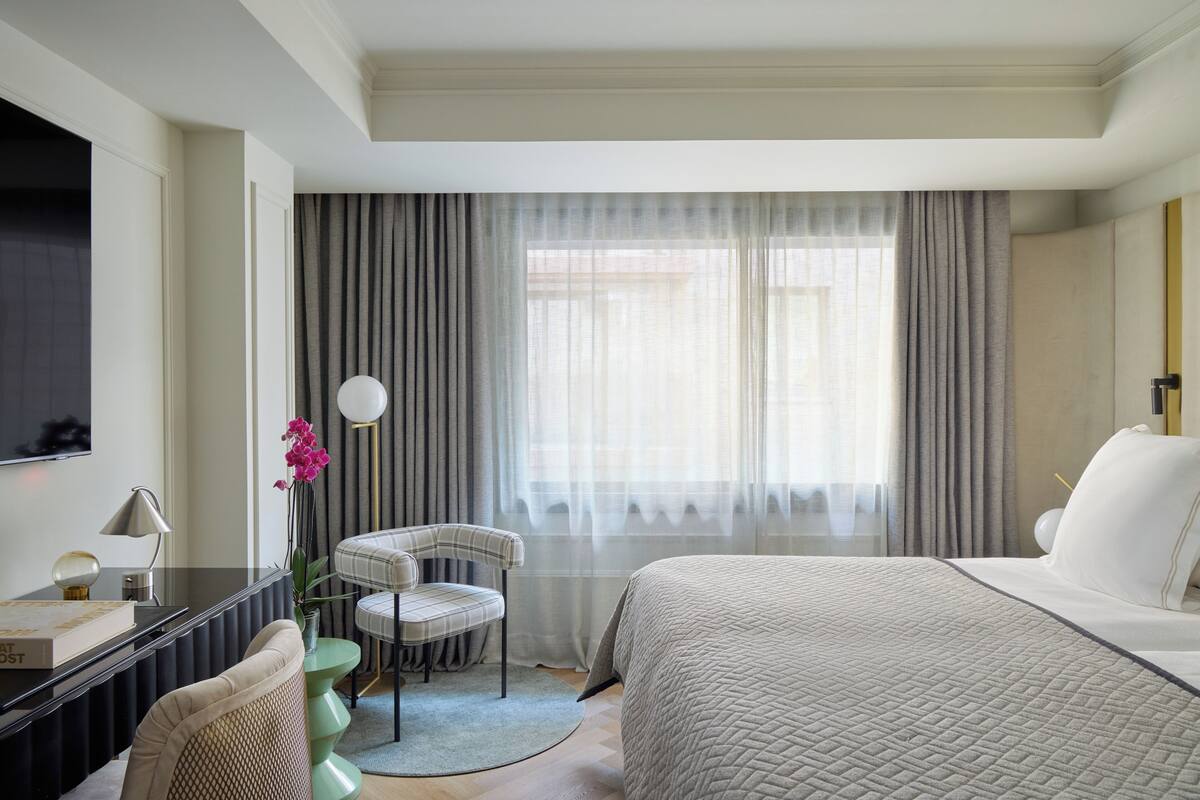 A hotel room with a bed, a desk with a lamp, a chair, a television, and a window with curtains.