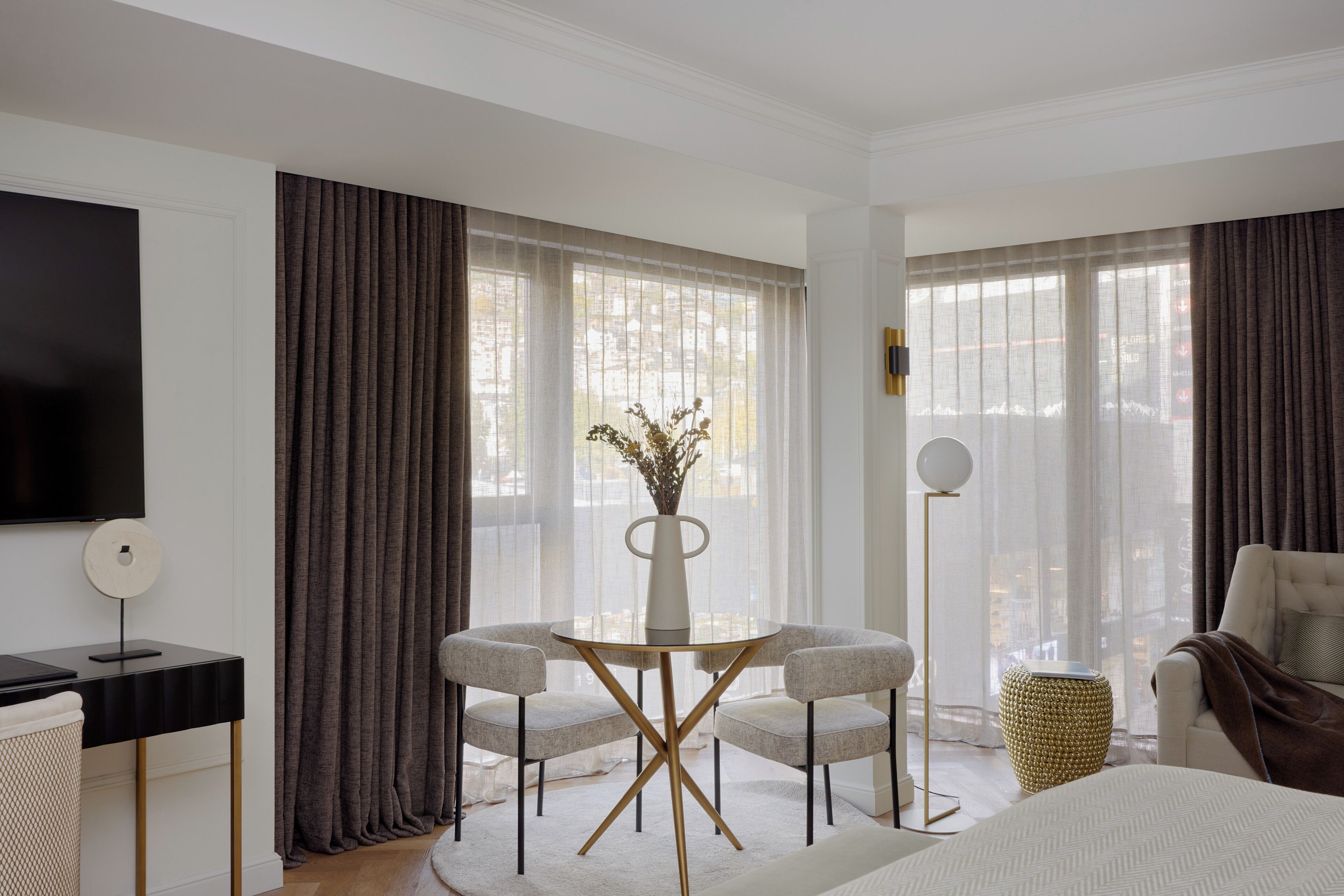 A hotel room with a bed, a desk with a TV, two armchairs, a round table with a vase, and large windows with curtains.
