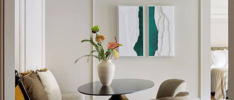 A modern living room with a round table, a sofa with cushions, and a vase with flowers. There is a framed artwork on the wall.