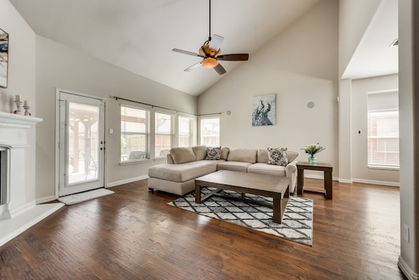A spacious living room with a sectional sofa, hardwood flooring, a ceiling fan, and a fireplace.