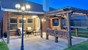 A patio area with a covered seating area, a pergola with string lights, and a brick house with a chimney. - New, beautiful 5 BR, 10 beds, near AT&T Stad,Joe Pool, Sixflag, and pet friendly (Grand Prairie)
