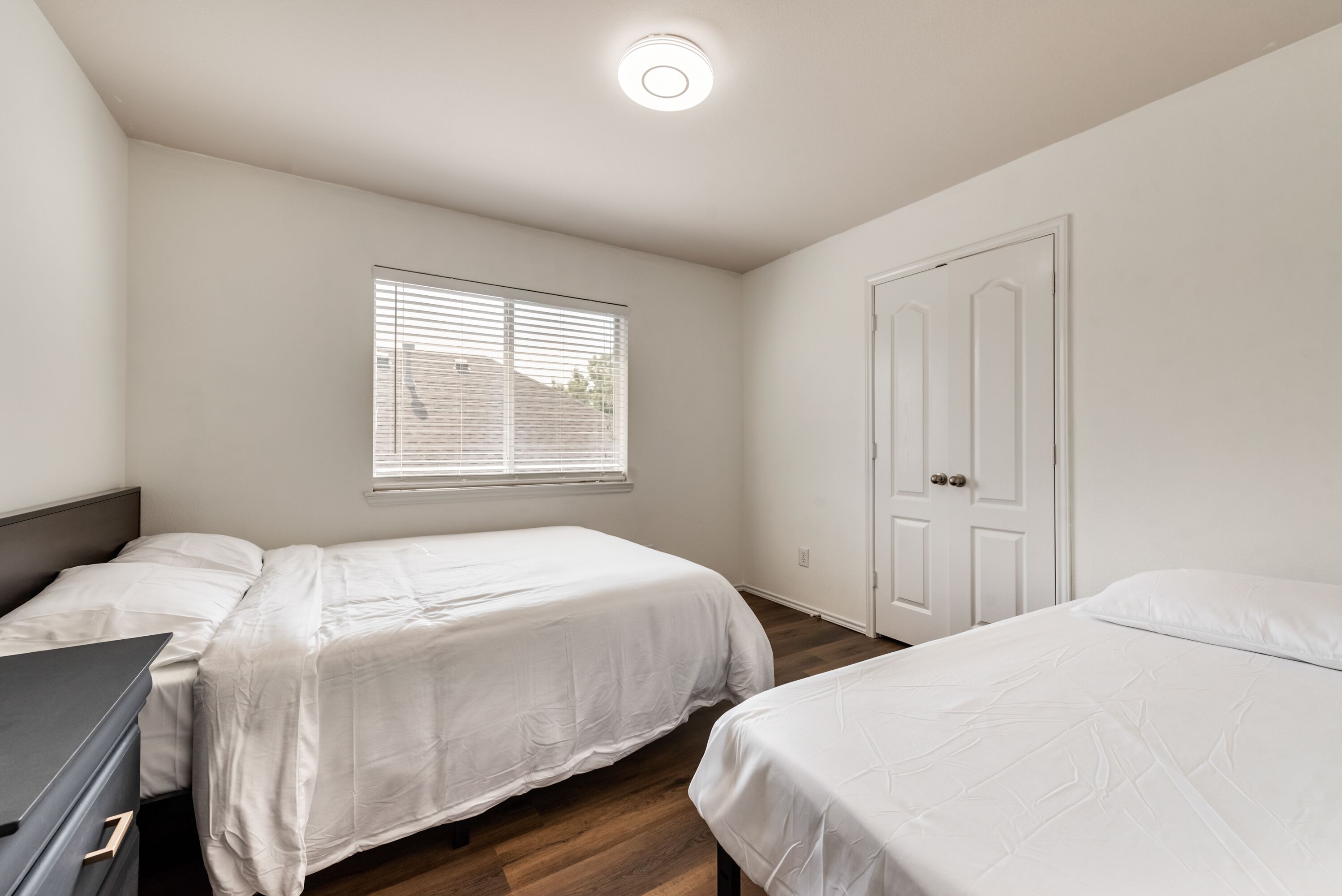 A bedroom with two beds, a dresser, a window with blinds, and a door.