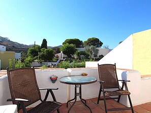 Une terrasse avec deux fauteuils en osier et une table ronde, donnant sur un jardin où poussent différentes plantes et offrant une vue lointaine sur des bâtiments.