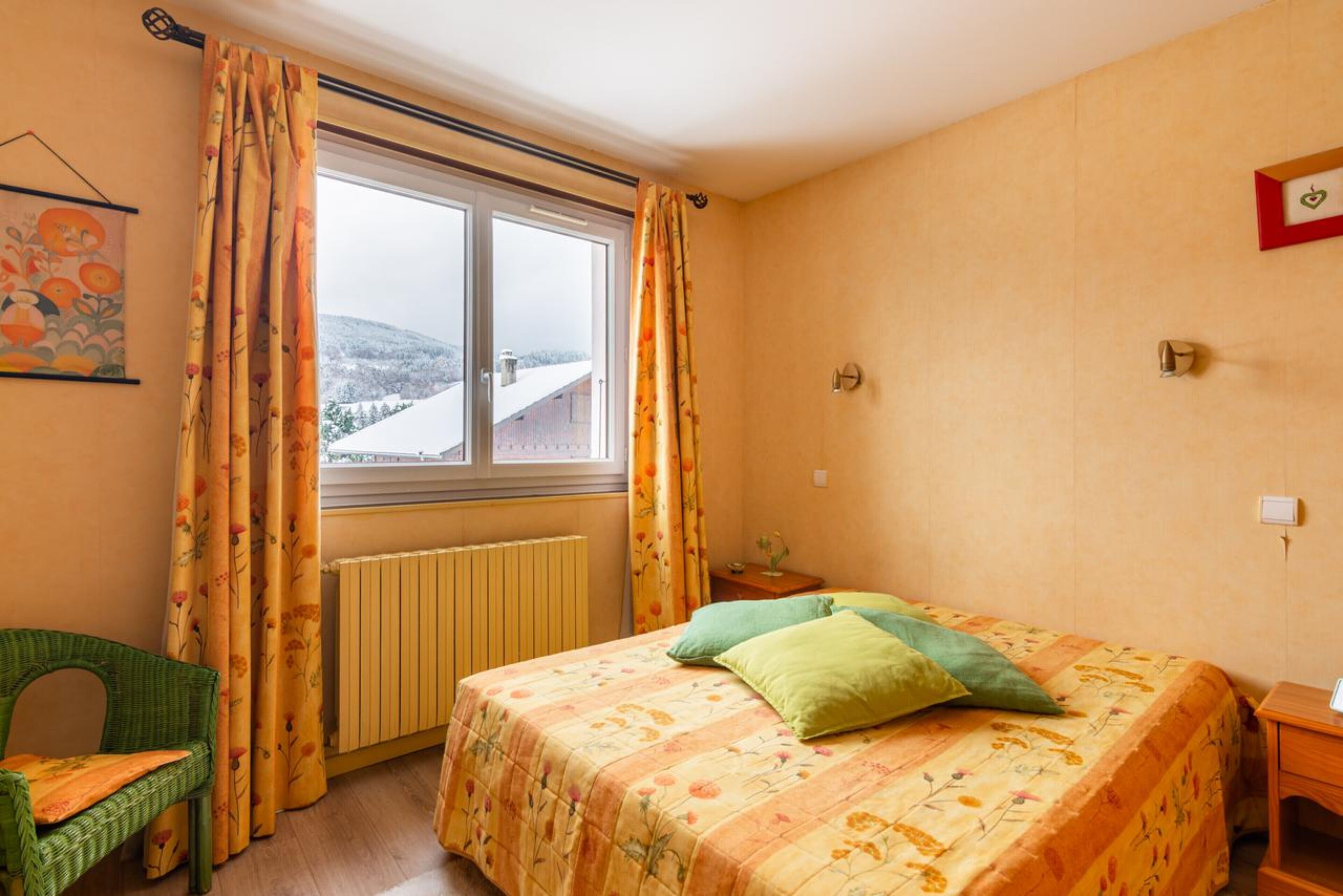 Un dormitorio con cama, una silla, una ventana con cortinas, un radiador y una lámpara fijada a la pared.