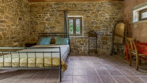 A stone-walled bedroom with a bed, a window, a mirror, and a red bench.