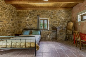 A stone-walled bedroom with a bed, a window, a mirror, and a red bench.