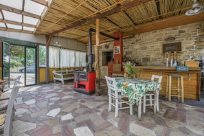 A rustic interior with a stone floor, wooden beams, a stone wall, a wooden table with a floral tablecloth, and a red stove.
