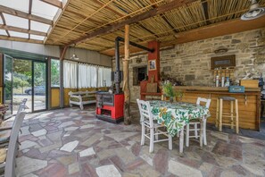 A rustic interior with a stone floor, wooden beams, a stone wall, a wooden table with a floral tablecloth, and a red stove.