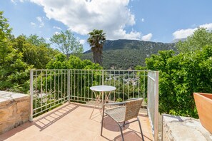 A balcony with a wicker chair and a small round table, overlooking a lush green landscape with a mountain in the background.