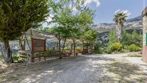 Un área de estar al aire libre, techada, con un sendero de grava, rodeada de árboles y montañas al fondo.