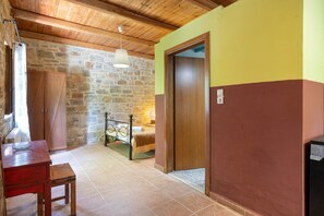 Una habitación con pared de piedra, techo de madera, una cama, un escritorio y una silla, y una puerta.