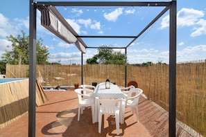 An outdoor patio with a table and chairs, a pergola, string lights, and a view of a pool and trees.