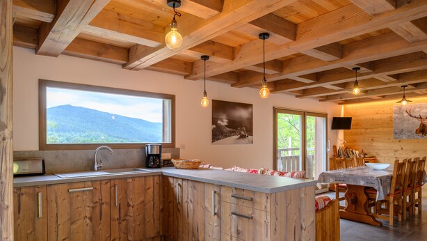Eine Küche mit Holzausstattung, einem großen Fenster, einem Essbereich mit Tisch und Stühlen sowie einem an der Wand befestigten Fernseher.