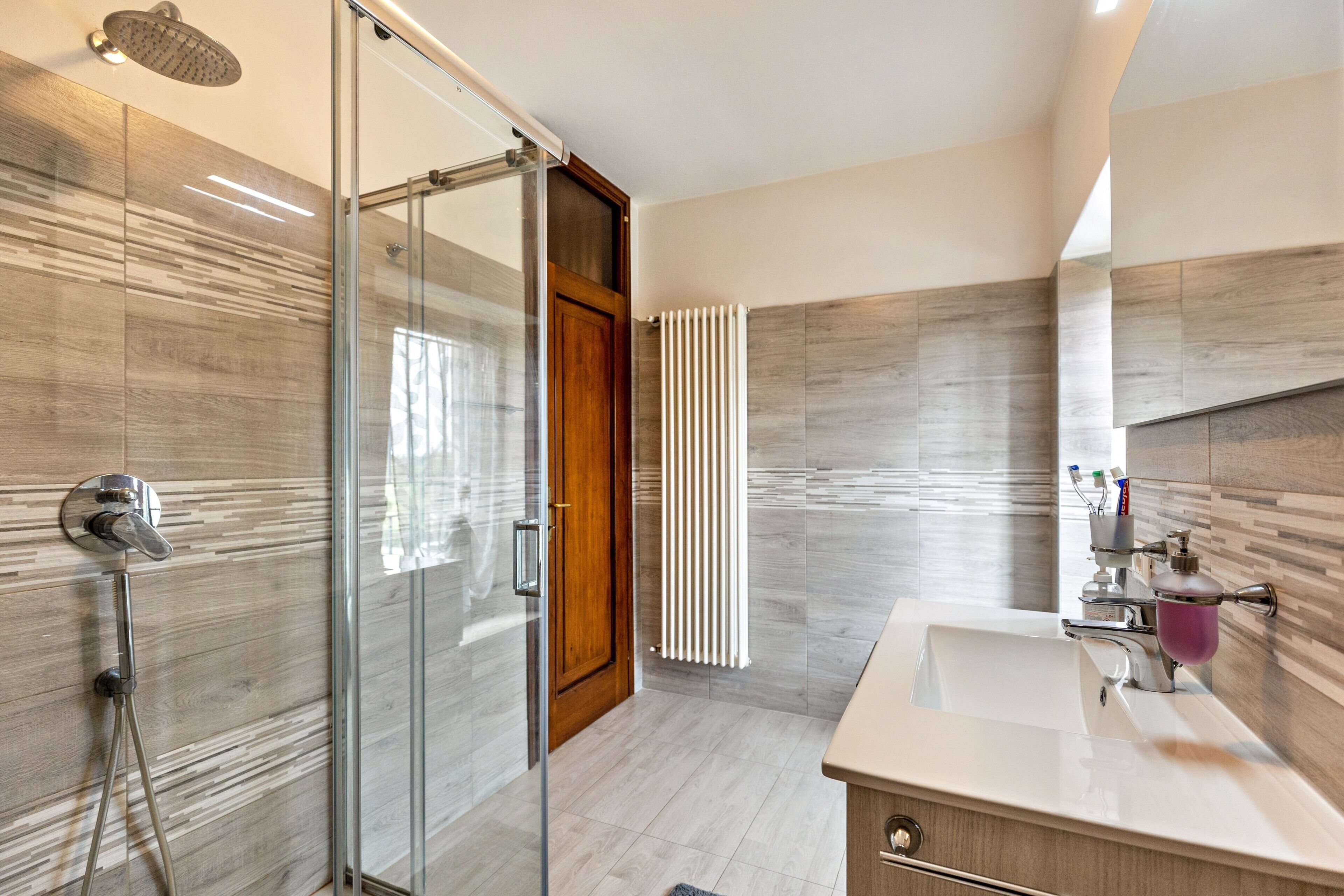 Een moderne badkamer met een douchecabine van glas, een houten deur, een witte wastafel en een spiegel.