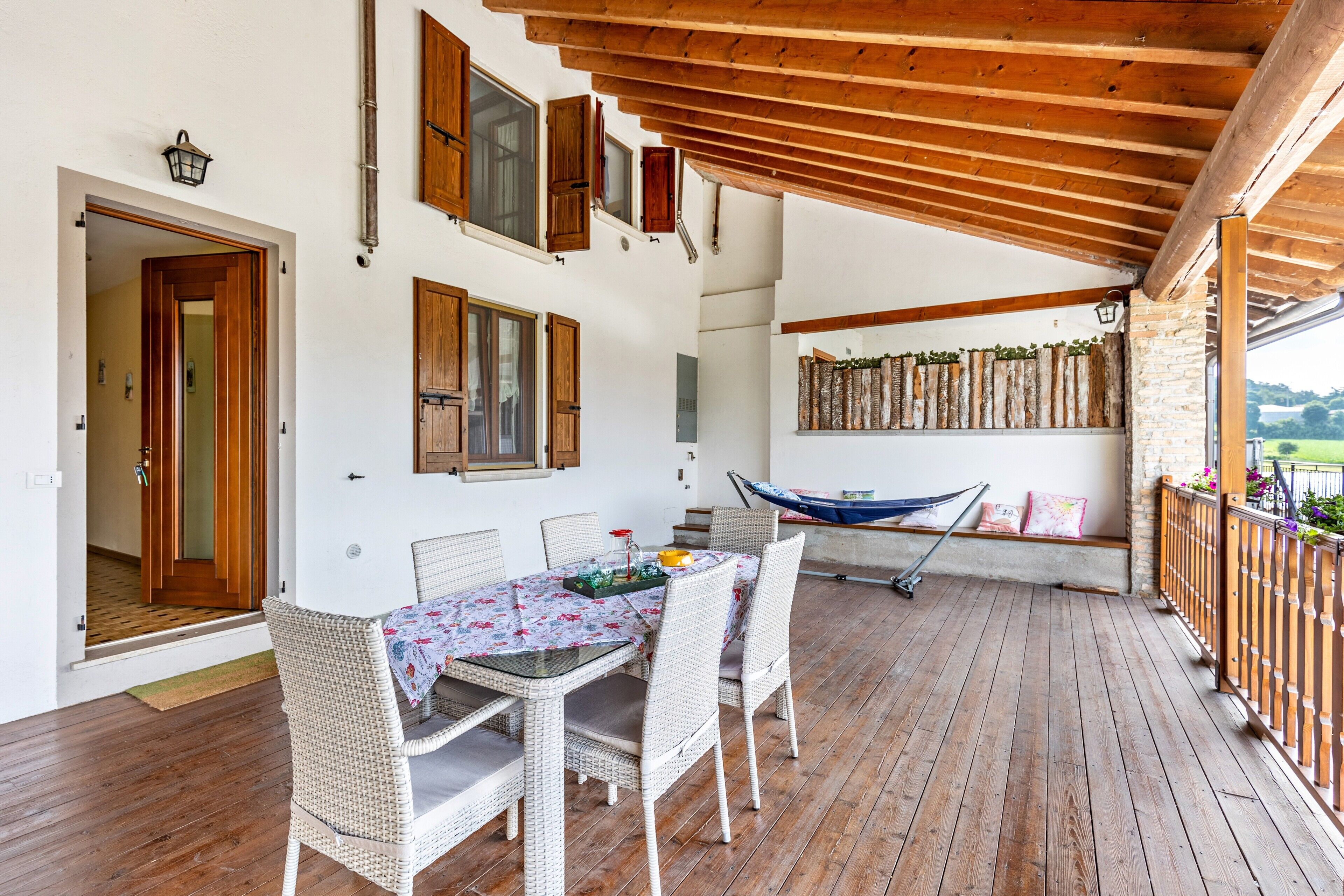 Une terrasse couverte avec une table en bois et des chaises en osier, un hamac et une vue sur l’extérieur.