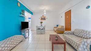 A living room with a sofa, a coffee table, a TV, a dining table, and a door. - Eftichia Nea Chora Apartment with Mountain View, Shared Garden & Wi-Fi (Chania)